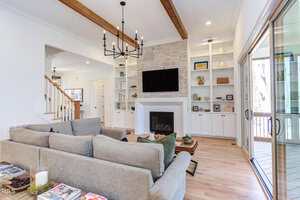 A modern living room with light wood floors, beige sectional sofa, a stone fireplace with a TV above it, built-in shelves, large windows, and exposed wooden ceiling beams. A staircase is visible to the left.