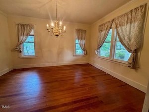 An empty room with hardwood floors, cream-colored walls, three windows with lace curtains, and a gold chandelier hanging from the ceiling. Natural light enters through the windows.