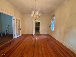 An empty room with wooden floors, cream-colored walls, a chandelier, a window with curtains, an open door, and double French doors leading to another room. A broom and cleaning supplies are visible in the adjacent space.