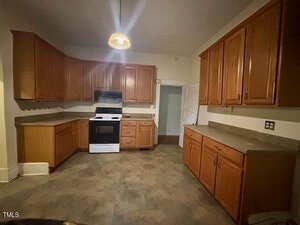 A kitchen with wooden cabinets, a white stove and oven, beige countertops, and grayish flooring. There is a single ceiling light and an open doorway in the back. The space appears empty and clean.