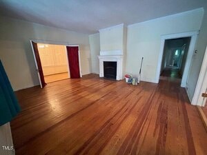 A mostly empty living room with hardwood floors, a white fireplace, and cleaning supplies on the floor. There are two doorways, one with red curtains leading to another room and one leading to a hallway.