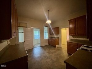 A dimly lit kitchen with brown cabinets, light countertops, and a hanging ceiling light. An open white door leads to a room with a window, and another doorway reveals a yellow-painted area.