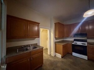 A kitchen with wooden cabinets, double sink, dark countertops, and a white gas stove with oven. There is a light fixture above and an open doorway leading to another room. The space is dimly lit.