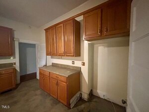 A kitchen with brown wooden cabinets, beige countertops, and an empty space likely for an appliance. The floor is gray, and there are visible outlets and some exposed cords on the lower right.