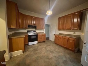 A kitchen with brown wooden cabinets, gray countertops, a white electric stove with a black range hood, a hanging light fixture, and neutral-colored flooring. There are two doors in the background.