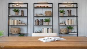 A wooden desk with an open book faces three black shelving units filled with books, plants, baskets, pottery, and decorative objects against a light striped wall. A beige chair is positioned at the desk.