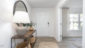 A bright entryway features a white front door, a wooden console table with a large lamp, a round mirror, decorative vases, woven baskets underneath, and light wood flooring. A window and curtains are visible to the right.