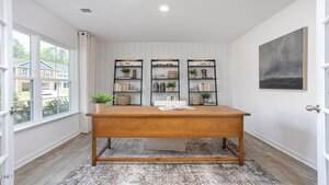 A home office with a large wooden desk, a patterned rug, two black bookshelves filled with books and decor, a potted plant, large windows, white walls, and a framed wall art piece.
