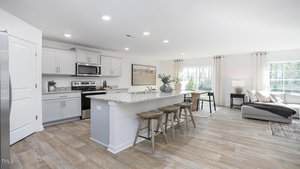 A modern, open-concept kitchen and living area with light gray cabinets, stainless steel appliances, a granite island with wooden stools, wood-look flooring, and a sectional sofa near a large window.