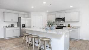 A modern kitchen with light gray cabinets, stainless steel appliances, granite countertops, and a large island with three wooden stools. Decorative vases with greenery are on the island. The floor is light wood.