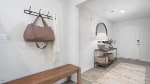 A modern entryway with a wooden bench, a wall hook holding a brown handbag and white scarf, a console table with a lamp, baskets underneath, a round mirror, and a white front door.