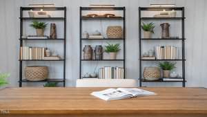 A wooden desk with an open book sits in front of three black bookshelves filled with neutral-toned decor, books, potted plants, woven baskets, and pottery against a light-colored wall.