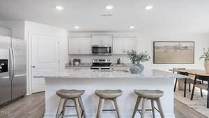 A modern kitchen with white cabinets, stainless steel appliances, and a granite island with three wooden stools. A dining table with chairs and a framed landscape picture are visible in the background.