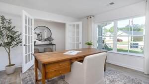 A bright home office with a wooden desk, beige upholstered chair, potted plant, and a large window letting in natural light. French doors lead to an adjacent room with a round mirror and console table.