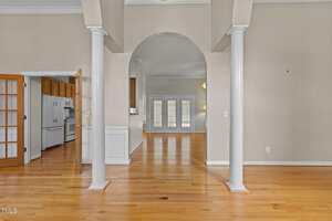 Open indoor space with light wood floors, beige walls, and white columns. An arched doorway leads to another room with double doors and shutters. A kitchen with wooden cabinets is visible to the left.