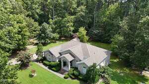 Aerial view of a single-story gray brick house with a gabled roof, surrounded by green lawn, shrubs, and dense trees under a clear sky. A covered entryway is visible at the front of the house.