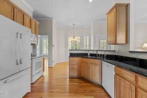 A kitchen with light wood cabinets, black granite countertops, white appliances, a double sink, hardwood floors, and a dining area with large windows and a chandelier in the background.