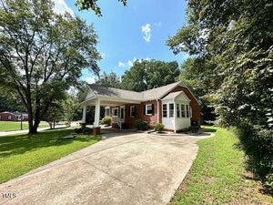 A one-story red brick house with white trim, a covered front porch, and a driveway. The house is surrounded by green grass, trees, and bushes under a blue sky with some clouds.
