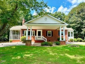 A single-story brick house with a covered front porch, white columns, and a small front stoop. There is a screened-in side porch and a well-kept lawn with trees and shrubs surrounding the property.
