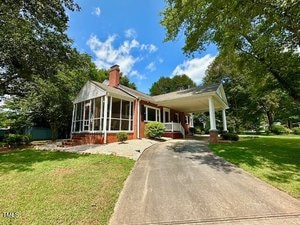 A single-story brick house at 1542 Oakdale Circle in Henderson features a screened porch, a carport with brick columns, a curved driveway, and a well-kept lawn surrounded by trees on a sunny day.
