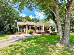 A single-story red brick house with white columns and trim, a front porch, a sloped driveway, and a large tree in the front yard, set against a background of green grass and leafy trees under a blue sky.