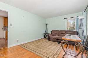 A living room with light green walls, a brown leather sofa, a patterned beige rug, two floor lamps, a glass-top table, and hardwood floors. A window with blinds lets in natural light.