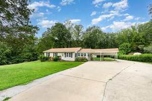 Single-story house with tan siding and a brown roof, featuring a covered carport and a wide concrete driveway. The yard is grassy, and the property is surrounded by trees under a partly cloudy sky.