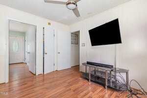 A room with hardwood floors, three white doors, a ceiling fan, a flat-screen TV mounted on the wall, a glass TV stand, and visible cables on the floor. A hallway with a decorative door is in the background.