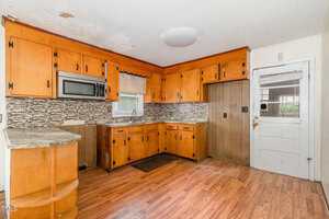A kitchen with wood cabinets, a microwave, a window above the sink, laminate flooring, a mosaic tile backsplash, and visible ceiling and wall damage. There is a white door with glass panels on the right.