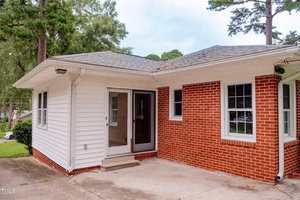 A one-story house with red brick and white siding exterior, featuring a glass storm door, white-framed windows, and a concrete patio area. The home is surrounded by trees and has a corner location.