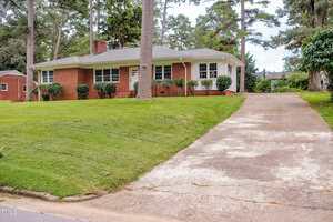 A single-story red brick house with white trim sits on a grassy, sloped lawn. A long concrete driveway leads up to the house, which is surrounded by tall trees and shrubs.