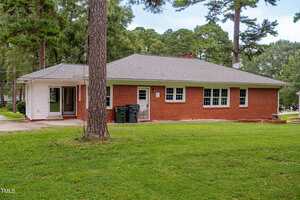 A single-story red brick house with a gray roof, multiple windows, and a white side extension. There are two garbage bins by the side wall and a grassy yard with several tall trees.
