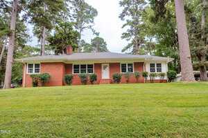 A single-story brick house with white trim at 105 W Thorndale Drive, Oxford, features large front windows, a gray shingled roof, and sits on a grassy lawn with shrubs surrounded by tall trees.
