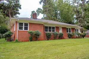 A single-story red brick house with multiple white-framed windows, a dark shingle roof, trimmed bushes in front, and a grassy lawn; surrounded by tall trees.