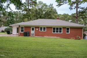 A single-story red brick house with multiple white-framed windows, a gray shingle roof, a side door, garbage bins, and a large grassy backyard surrounded by trees.