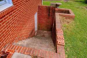 Outdoor concrete steps with red brick walls lead down to a basement door. Loose bricks are stacked on the right wall, and there is a patch of grass next to the stairwell. The brick exterior belongs to a house.