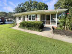 Charming single-story home at 613 15th Street, Butner, featuring beige siding with white trim, a screened front door, multiple windows, concrete walkway, trimmed hedges, and a well-maintained lawn under a blue sky with scattered clouds.