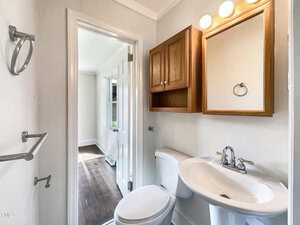 A small bathroom with a white toilet, pedestal sink, and a wooden mirrored cabinet above the sink. The walls are white, and the bathroom opens into a room with wood flooring and natural light.