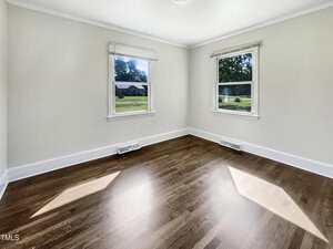 Empty room with light-colored walls, two windows with white trim, hardwood floors, white baseboards, and two floor vents beneath the windows. Sunlight enters through the windows, casting bright patches on the floor.