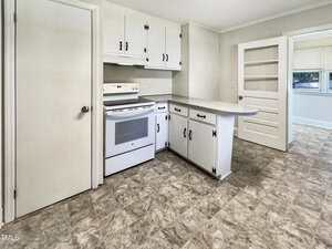 A kitchen with white cabinets, a white electric stove, and a small peninsula counter. The floor is patterned vinyl, and there are doors leading to other rooms and a set of built-in shelves.