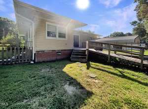 Single-story beige house with a brick foundation, wooden ramp, steps, and fenced area. The sun is bright in a clear blue sky, casting shadows on the grassy yard. Other homes and trees are in the background.