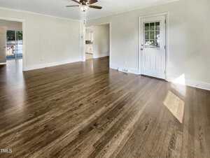 A spacious, empty room with hardwood floors, light-colored walls, a ceiling fan, a white door with a window, and an open entryway leading to a bright kitchen area. Natural light enters through the door's window.