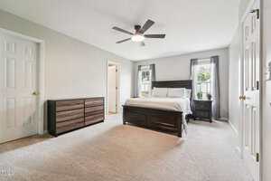 A spacious bedroom with light carpet, a dark wood bed frame, matching dresser, two windows with gray curtains, a ceiling fan, and neutral-colored walls. A white door and closet door are visible.