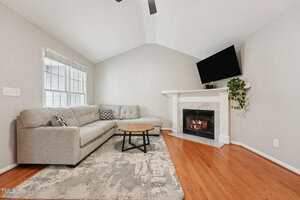A living room with a gray sectional sofa, a round wooden coffee table, a fireplace with a TV mounted above it, a potted plant, large windows, light gray walls, and hardwood floors.