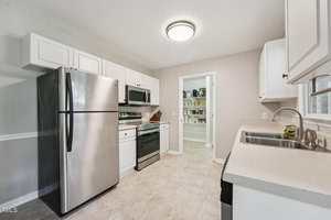 A modern kitchen with white cabinets, stainless steel appliances including a refrigerator, oven, and microwave, light countertops, and a view into a pantry with shelves. The room is well-lit and has a clean, neutral design.