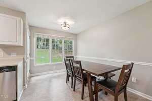 A dining area with a dark wooden table and four matching chairs, neutral-colored walls, a large window with white trim, and a modern ceiling light fixture. Natural light fills the space.