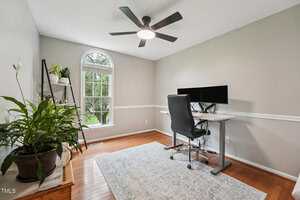 A home office with a large window, an adjustable desk with dual monitors, a black office chair, several potted plants, light-colored walls, and hardwood floors covered partially by a rug.