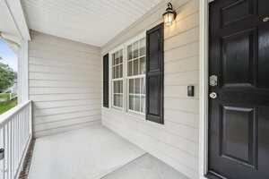 Covered front porch with light beige siding, a double window with black shutters, a black front door, a wall-mounted light fixture, concrete floor, and white railing.