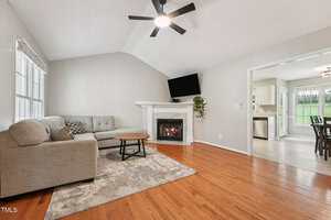 A living room with a sectional sofa, an area rug, a round coffee table, a fireplace with a TV above it, wood floors, and an open view to a dining area and kitchen with tile flooring.