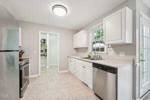 A modern kitchen with white cabinets, stainless steel appliances, a single basin sink, and a window above the sink. There is a door leading outside and an open pantry visible through a doorway.